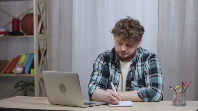 穿着方格衬衫的年轻人看着笔记本电脑屏幕无聊，在笔记本电脑上做笔记。留着胡子的红发男性坐在家庭办公室的