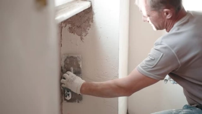 工作服修理工用金属抹刀、抹刀和白色抹灰糊将破旧的水泥墙放在窗下，用腻子粉刷平整墙面。墙壁修复，大修概