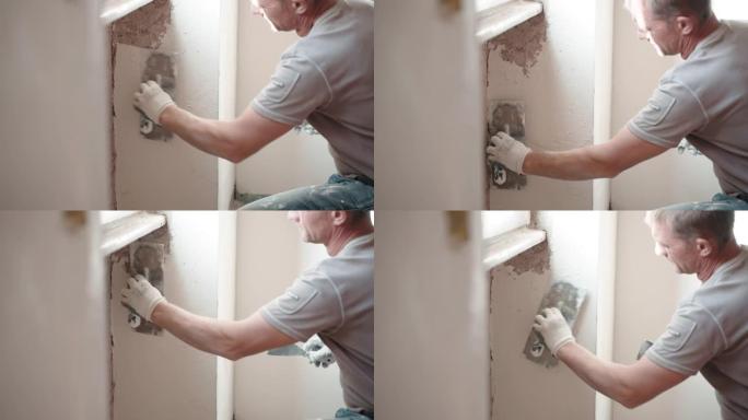 工作服修理工用金属抹刀、抹刀和白色抹灰糊将破旧的水泥墙放在窗下，用腻子粉刷平整墙面。墙壁修复，大修概