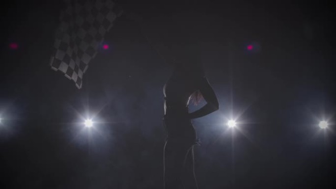 剪影的侧视图年轻女子挥舞着方格竞赛旗，标志着赛车活动的开始。黑发女人在背光的黑暗烟熏工作室摆姿势。特