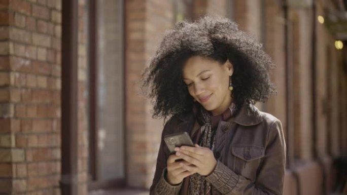 年轻的非洲裔美国妇女在智能手机上发短信的肖像。穿着皮夹克的黑发女人在模糊的砖砌建筑的背景下走在街上。