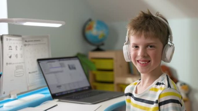 戴着耳机的七年金发男孩笑了。男孩坐在家里的笔记本电脑上。在线课程。