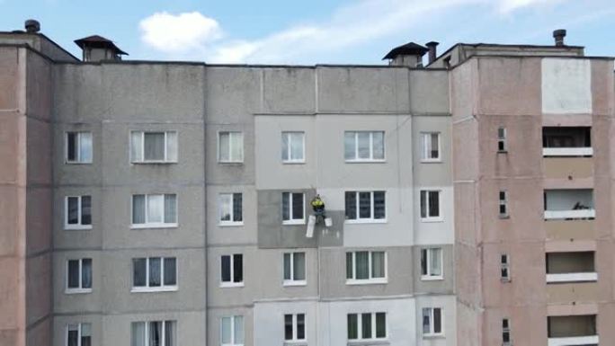 一个工业登山者悬挂在绳索上，用滚筒在建筑物的墙壁上粉刷。一名男性工人在多层建筑物的外墙上进行隔热工作