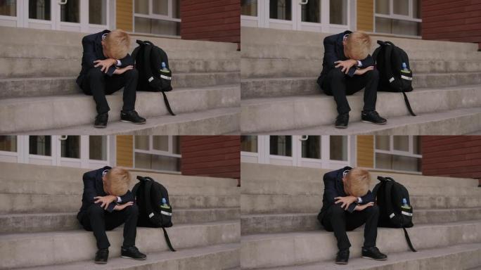 一个悲伤的男孩坐在学校楼梯上哭泣，他的背包就在他旁边。