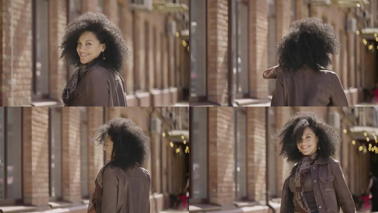 年轻的非洲裔美国妇女走在街上，转身微笑。穿着棕色皮夹克的黑发女人在模糊的砖砌建筑中摆姿势。特写。慢动