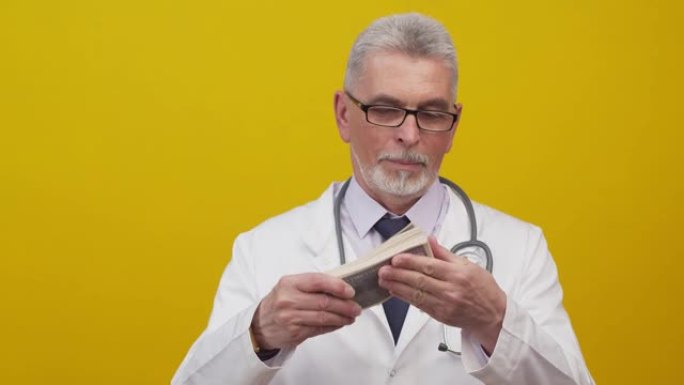 成年医生穿着医学礼服和带有听诊器的眼镜挥舞着一捆美元钞票，并在工作室的橙色背景上迅速叙述。有偿用药的