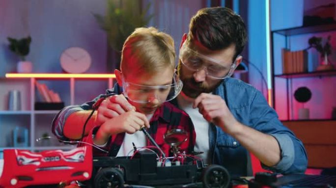 好看的满意的有爱心的成人胡子的父亲在防护眼镜上教焊接玩具车的小细节他好奇聪明的少年儿子也戴防护眼镜