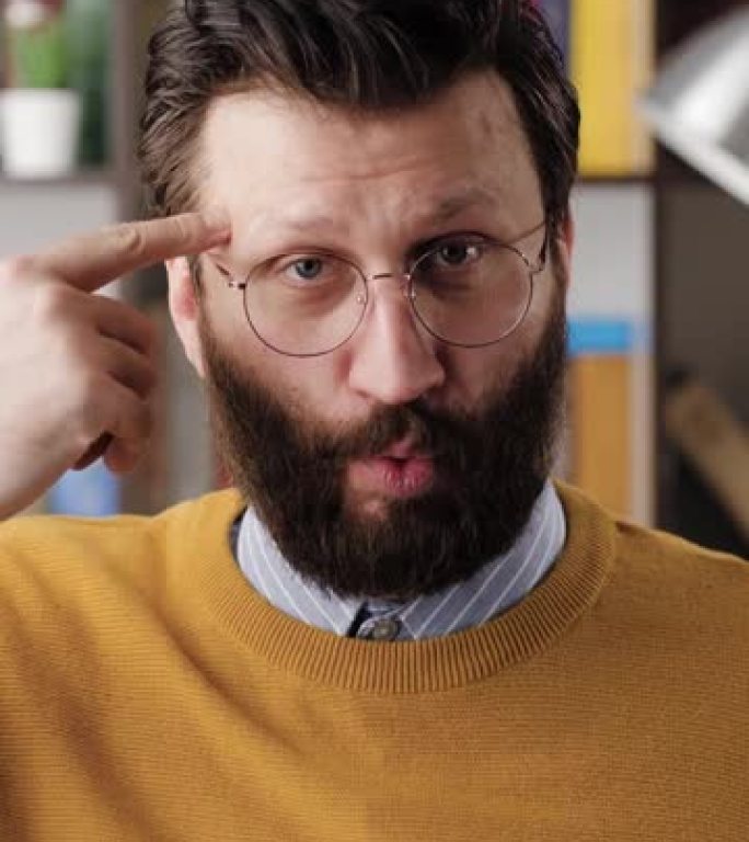 愚蠢。办公室或公寓房间里戴着眼镜的大胡子男人的垂直视频看着相机，手指在太阳穴上旋转