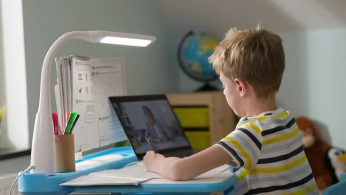 男孩坐在家里的笔记本电脑上。在线课程。孩子一边看笔记本电脑屏幕，一边写笔记本电脑。