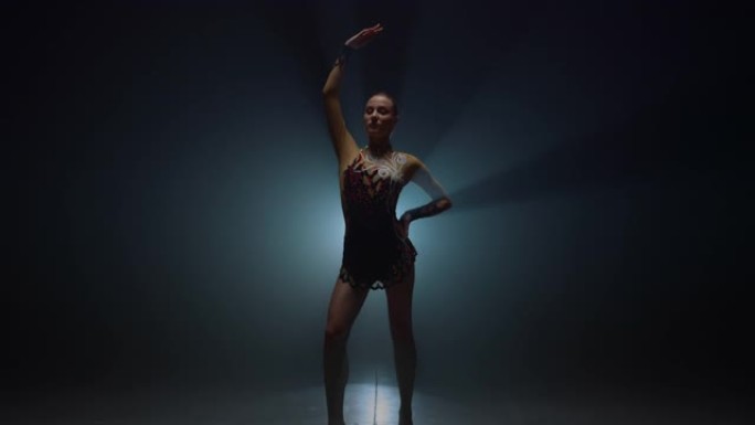 体操女孩举起手站在黑暗的空间。女运动员在室内摆姿势。