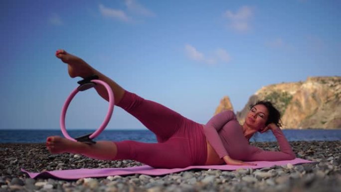一个黑发的年轻女子在圆石滩海边的瑜伽垫上做普拉提。女性健身瑜伽常规概念。健康的生活方式，和谐与冥想