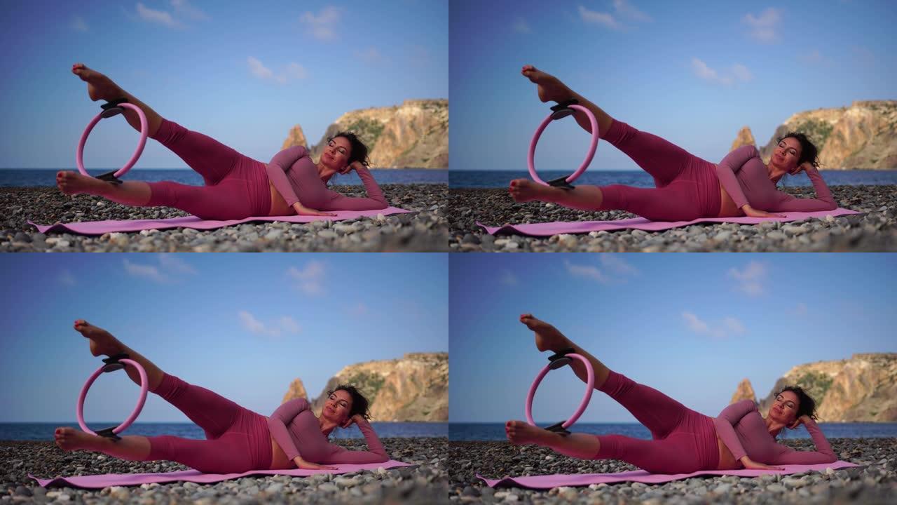 一个黑发的年轻女子在圆石滩海边的瑜伽垫上做普拉提。女性健身瑜伽常规概念。健康的生活方式，和谐与冥想