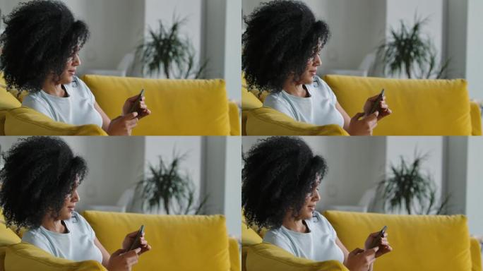 一位年轻的非洲裔美国妇女在手机上发短信的肖像。黑发，卷发，坐在明亮的家庭房间里的黄色沙发上。特写。慢