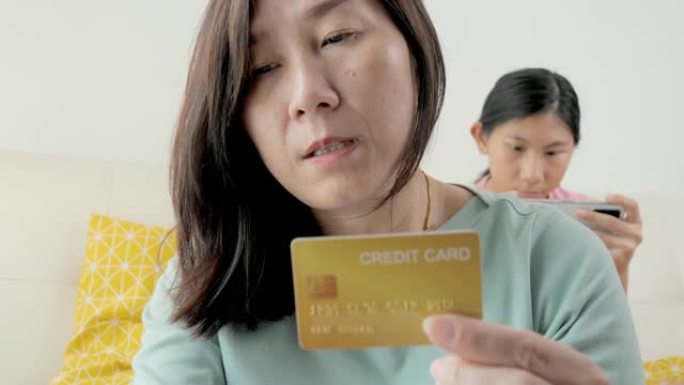 亚洲母亲使用信用卡进行在线购物或结账，而女儿则在家中沙发上放松，这是生活方式的概念。
