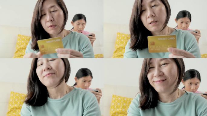 亚洲母亲使用信用卡进行在线购物或结账，而女儿则在家中沙发上放松，这是生活方式的概念。