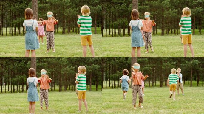 一群孩子在公园玩盲人的buff标签游戏。蒙住眼睛的小女孩试图触摸朋友拍手并跑来跑去。孩子们在暑假玩得