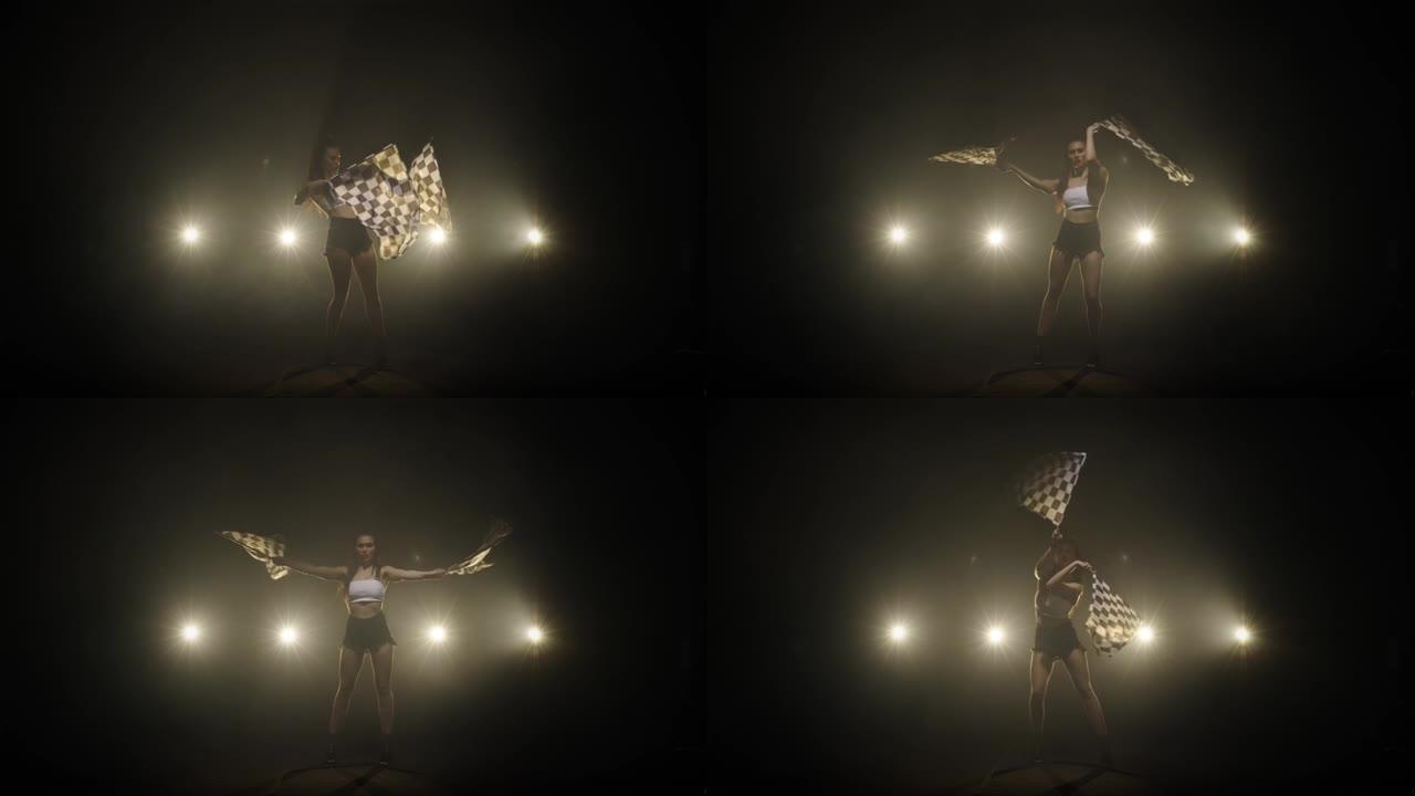 漂亮的年轻女子在赛车比赛中表演时挥舞着方格种族旗帜。深色皮肤的剪影在一个带背光的黑暗烟熏工作室中呈现