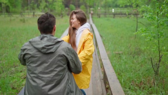 浪漫的年轻夫妇在乡村的木路上慢动作奔跑。快乐的爱心女人牵着男朋友的手，男人在雨中跟着她去森林