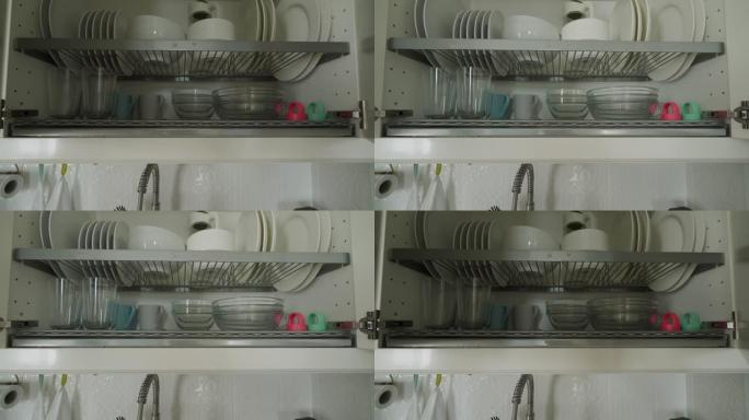 男人打开厨房用干净的餐具关闭橱柜，白色的盘子和杯子在厨房里晾干。