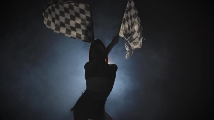 苗条的年轻女子挥舞着方格竞赛旗的剪影，标志着赛车活动的开始。黑发女人在一个带背光的黑烟工作室摆姿势。