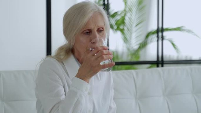 老年妇女坐在沙发上，用玻璃杯喝水