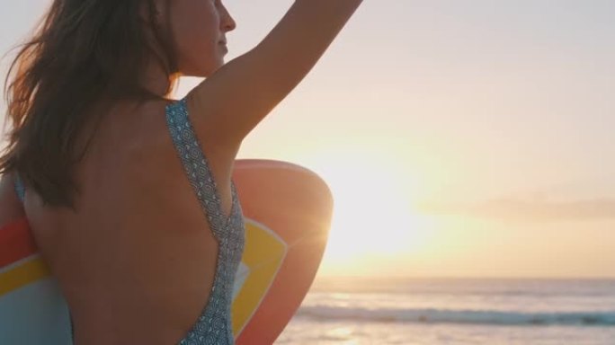 女冲浪者。年轻女子站在沙滩上冲浪板上，看着日出时的海浪