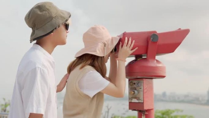 欢快的亚洲年轻夫妇游客在观看点使用投币式双筒望远镜观看城市背景。年轻人的关系。