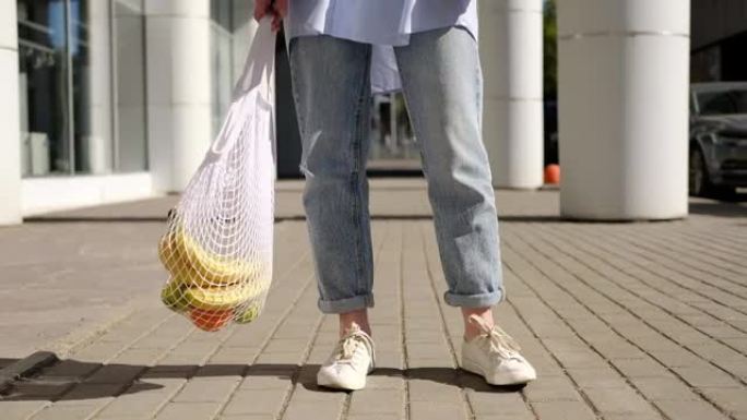 穿着牛仔裤的女人拿着，摇晃着，摇晃着白色网眼袋。环保、可重复使用的购物袋。橘子、苹果、香蕉装在棉针织