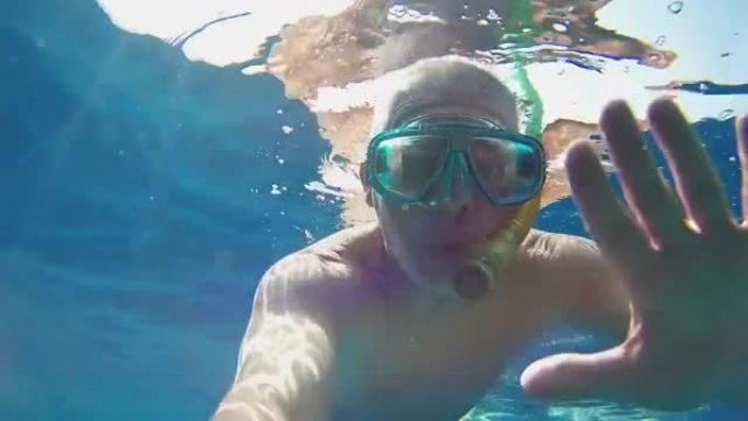 一个戴着面具和通气管的男人在海水下挥舞着手，向Victoria GoPro展示手指