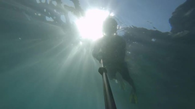 一个戴着面具和通气管的年轻运动男子在水下深处用自拍杆游泳。阳光透过水