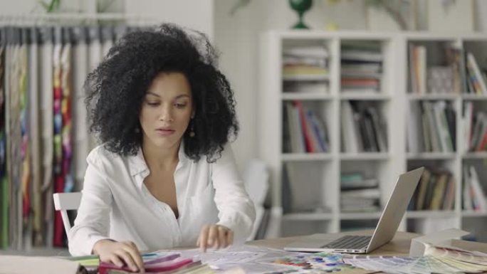 年轻的非洲裔美国妇女的肖像，通过调色板和面料样本寻找设计项目。女设计师坐在办公室的桌子旁。特写。慢动