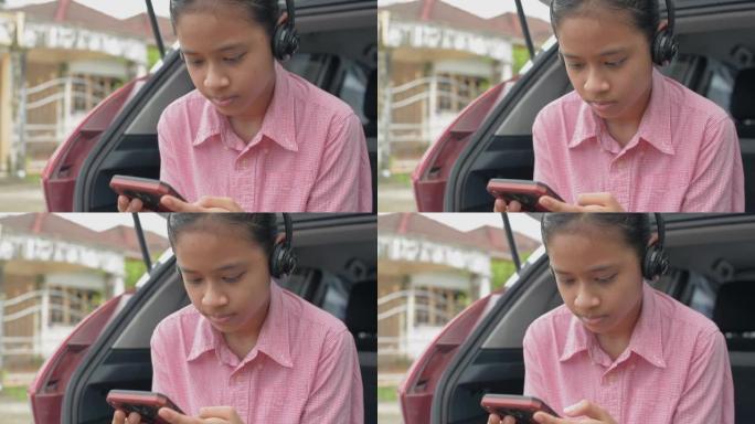 亚洲女少年观看社交媒体或通过带耳机的智能手机在线玩游戏，并坐在掀背车的后侧，同时等待父母开车送她上学