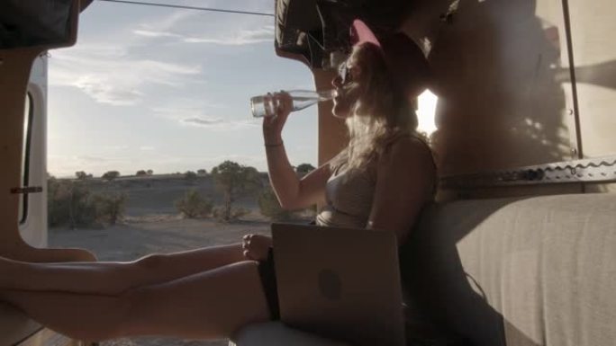 夏季，黄昏时分，科罗拉多州弗鲁伊塔附近，穿着软呢帽的千禧一代金发女子坐在露营车的后面喝瓶装水