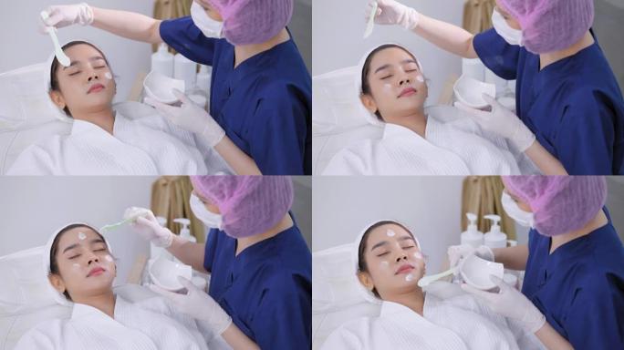 戴着卫生发帽的医务人员为年轻的亚洲妇女提供面部护理服务，她穿着浴袍躺在白色的医疗沙发上，在眼部接受保