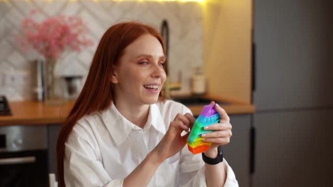 快乐放松的红发年轻女子推着彩色彩虹色柔软的硅胶泡泡坐在轻便厨房的桌子旁。