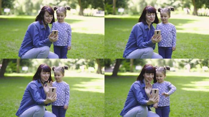 妈妈和可爱的3岁小女儿，穿着休闲的蓝色衣服，用手机摄像头自拍，给朋友打视频，挥手，在阳光明媚的夏日公