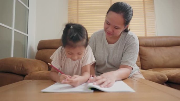 亚洲母亲照顾她的小女孩在家学习学校练习册上的写作