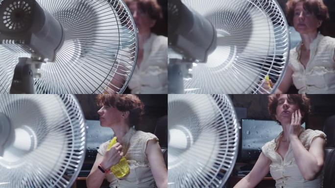 女人正试图在家里的台式计算机上工作，遭受炎热的天气并在使用电风扇时向她的脸上喷水
