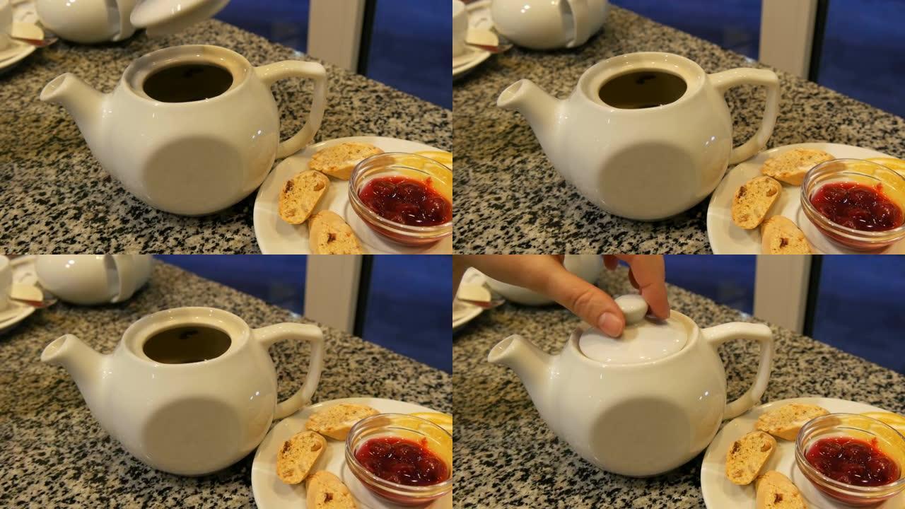 女人的手抬起白色茶壶的盖子。陶瓷白茶壶和茶托茶具，旁边是饼干，柠檬片和浆果果酱