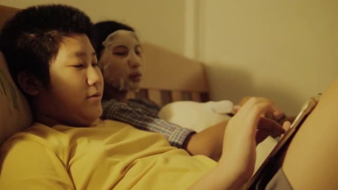 亚洲儿童晚上在家一起在床上用数字平板电脑看电影，女孩用面膜遮住脸，生活方式理念。