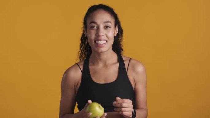 年轻漂亮的非洲裔美国妇女穿着运动胸罩，拿着苹果，在黄色背景下训练后看起来很开心。运动女孩