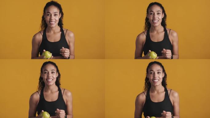 年轻漂亮的非洲裔美国妇女穿着运动胸罩，拿着苹果，在黄色背景下训练后看起来很开心。运动女孩