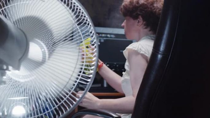 女人正试图在家里的台式计算机上工作，遭受炎热的天气并在使用电风扇时向她的脸上喷水
