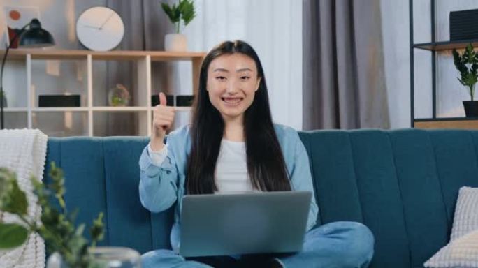 好看的自信微笑的25岁亚洲女孩，穿着便装的黑色长发，坐在沙发上，指着相机的手势