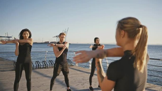 一群不同的人在户外锻炼，在健身教练后重复热身运动，一起站在码头上