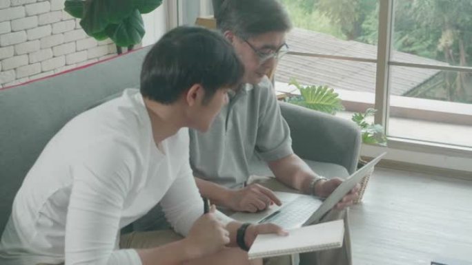 带着儿子的家庭坐在沙发上看笔记本电脑，并在记事本上写清单费用财务与在家中的高级父亲，老人和年轻人在线
