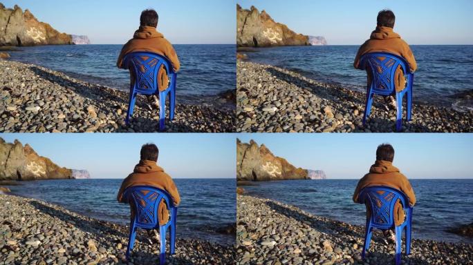 一名成年男性匿名者坐在春秋卵石滩上的蓝色椅子上欣赏海景的后视图。淡季旅行