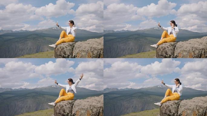 女游客坐在山崖边的智能手机相机上自拍。旅行者视频通话并与朋友分享他的印象