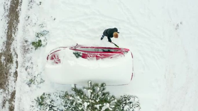 年轻女子早上在停车场自上而下的视野中清理一辆冰雪覆盖的汽车。美丽的女人在寒冷的冬天早晨刷她的雪车。
