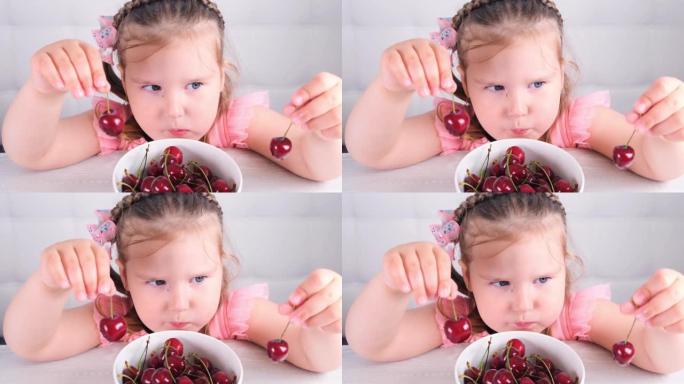 可爱的小女孩坐在一张轻便的木桌旁，桌上放着一盘樱桃，手里拿着红色浆果。健康饮食